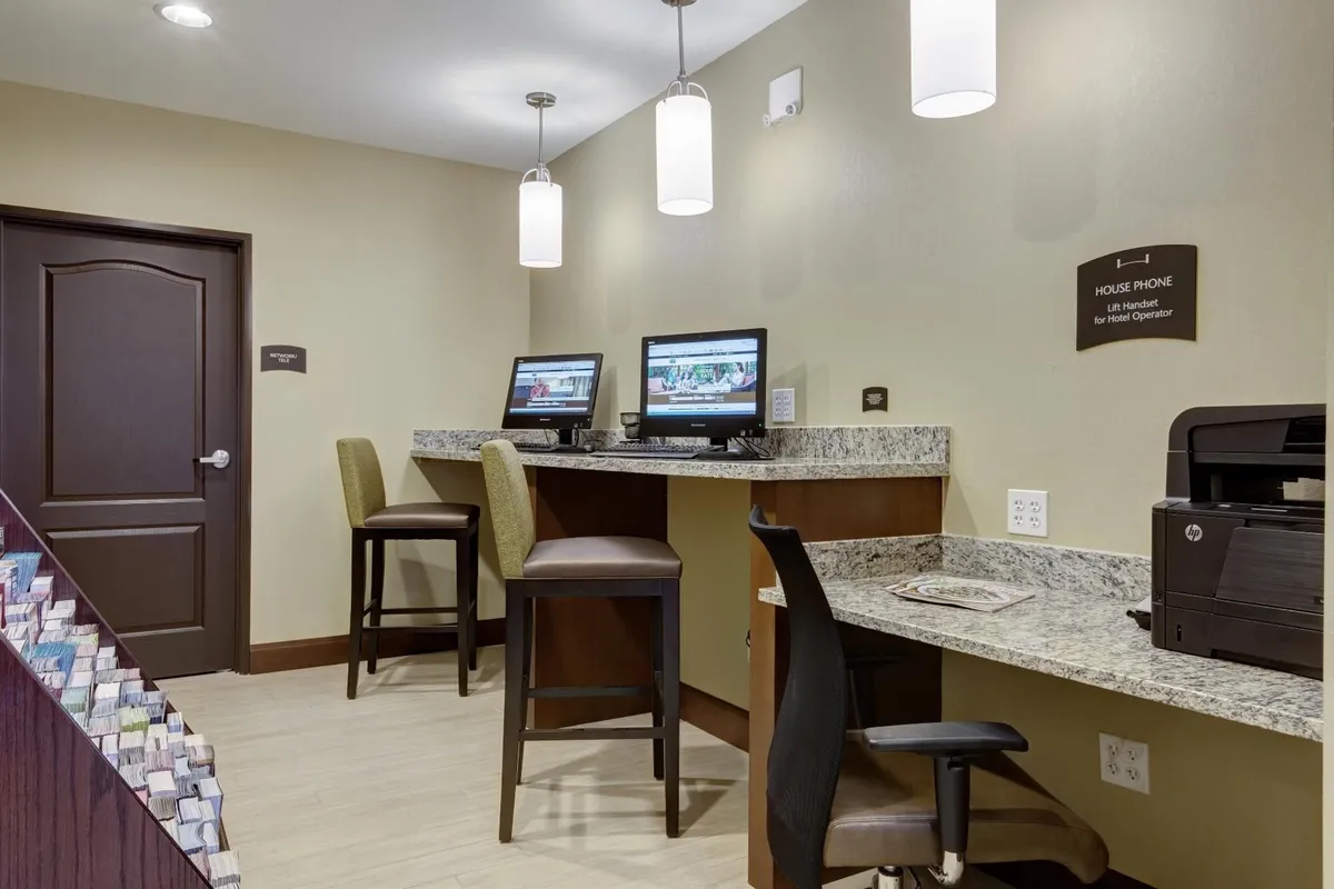 This interior image shows a hotel business center with computers on a granite countertop, bar stools for seating, and a house phone available for guest use. The atmosphere is practical and inviting, with friendly staff assistance evident and easy access to technology for guests needing to catch up on work or travel planning.