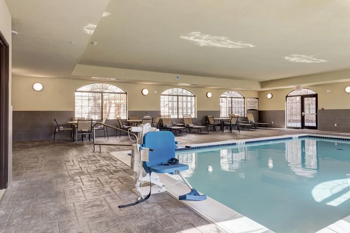 This interior image showcases a calm and inviting indoor swimming pool area with poolside seating and large windows allowing plenty of sunlight to reflect on the water. The atmosphere appears peaceful, offering opportunities for relaxation and quiet moments, with a touch of natural light enhancing the tranquil mood.