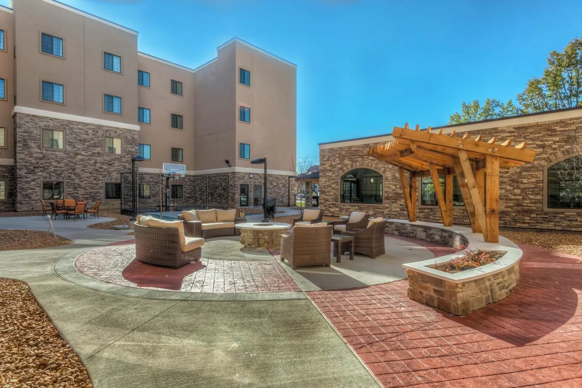 An inviting outdoor lounge area featuring a fire pit surrounded by comfortable cushioned seating beneath a wooden pergola. Brick pathways weave through the landscaped exterior, perfect for relaxing evenings and social gatherings. The setting is cozy and ideal for family conversations and unwinding after a day of adventure.