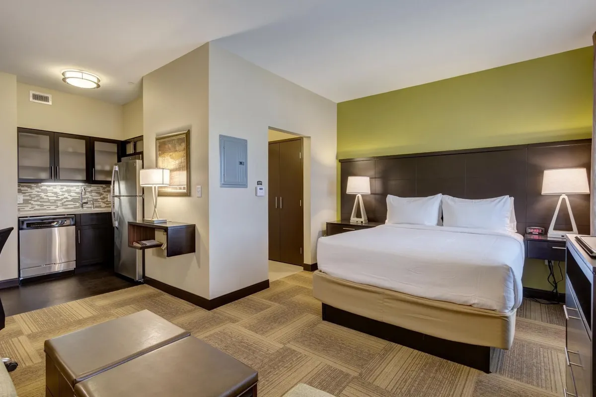 The image showcases a bright, spacious studio with a plush king-sized bed, a modern kitchen area equipped for meal prep, and a dedicated workspace with a comfortable chair—all seamlessly integrated into an inviting open layout.