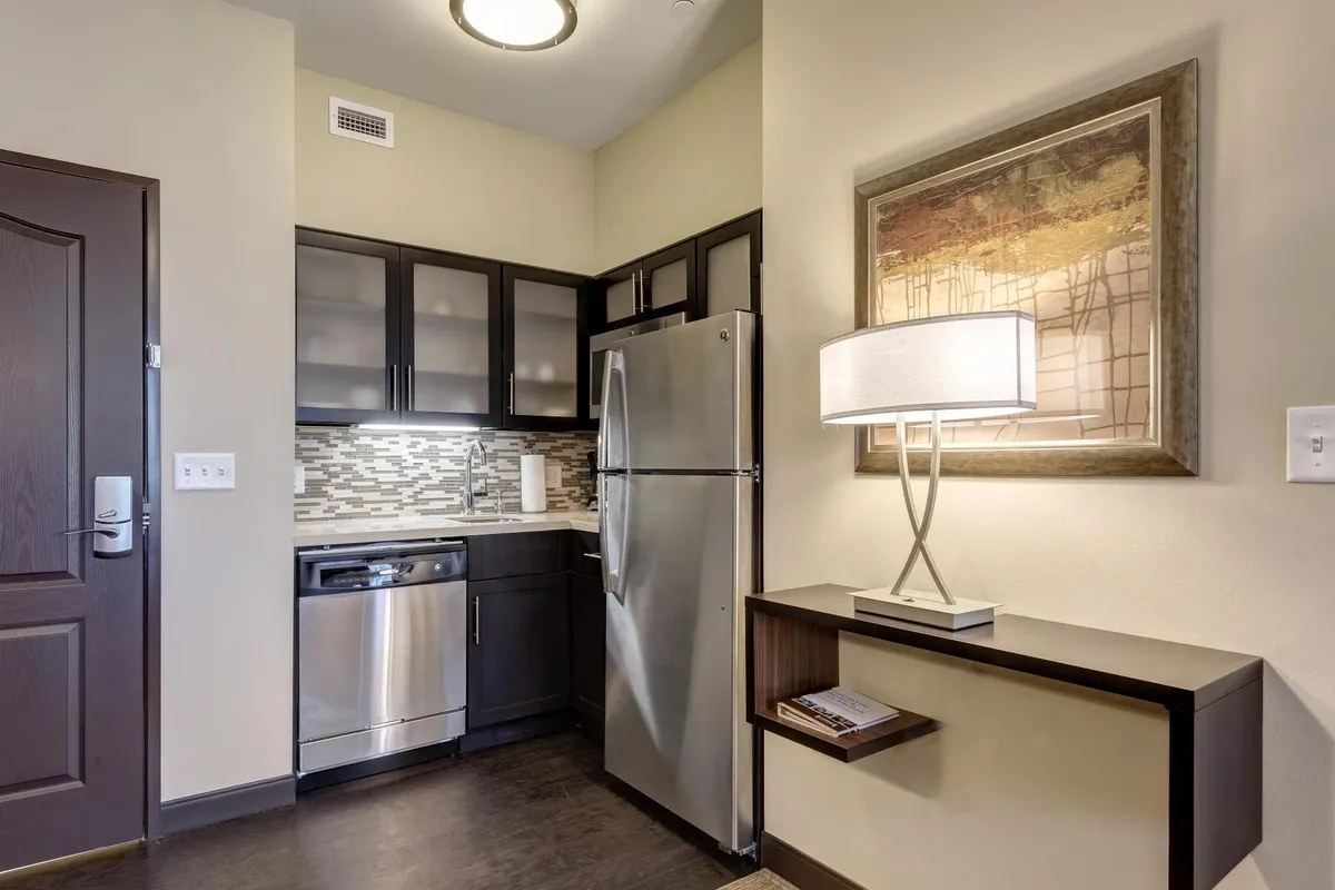 A modern kitchen and living area featuring sleek cabinetry, kitchen appliances, and tasteful artwork on the wall. The space is cozy and contemporary, suggesting a comfortable and stylish atmosphere suited for relaxation or entertaining. Visible activities include preparing a meal, sharing drinks, and enjoying coffee, all set within a well-appointed studio suite environment.