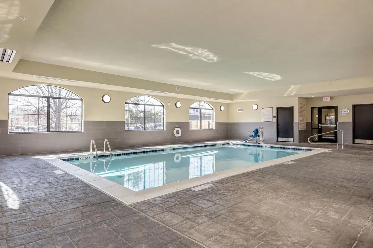 A serene indoor swimming pool with expansive windows allowing in natural light. The water sparkles under the sunlight, and comfortable lounge seating lines the poolside. The scene captures a calm moment, with guests enjoying a dip or simply relaxing by the water.